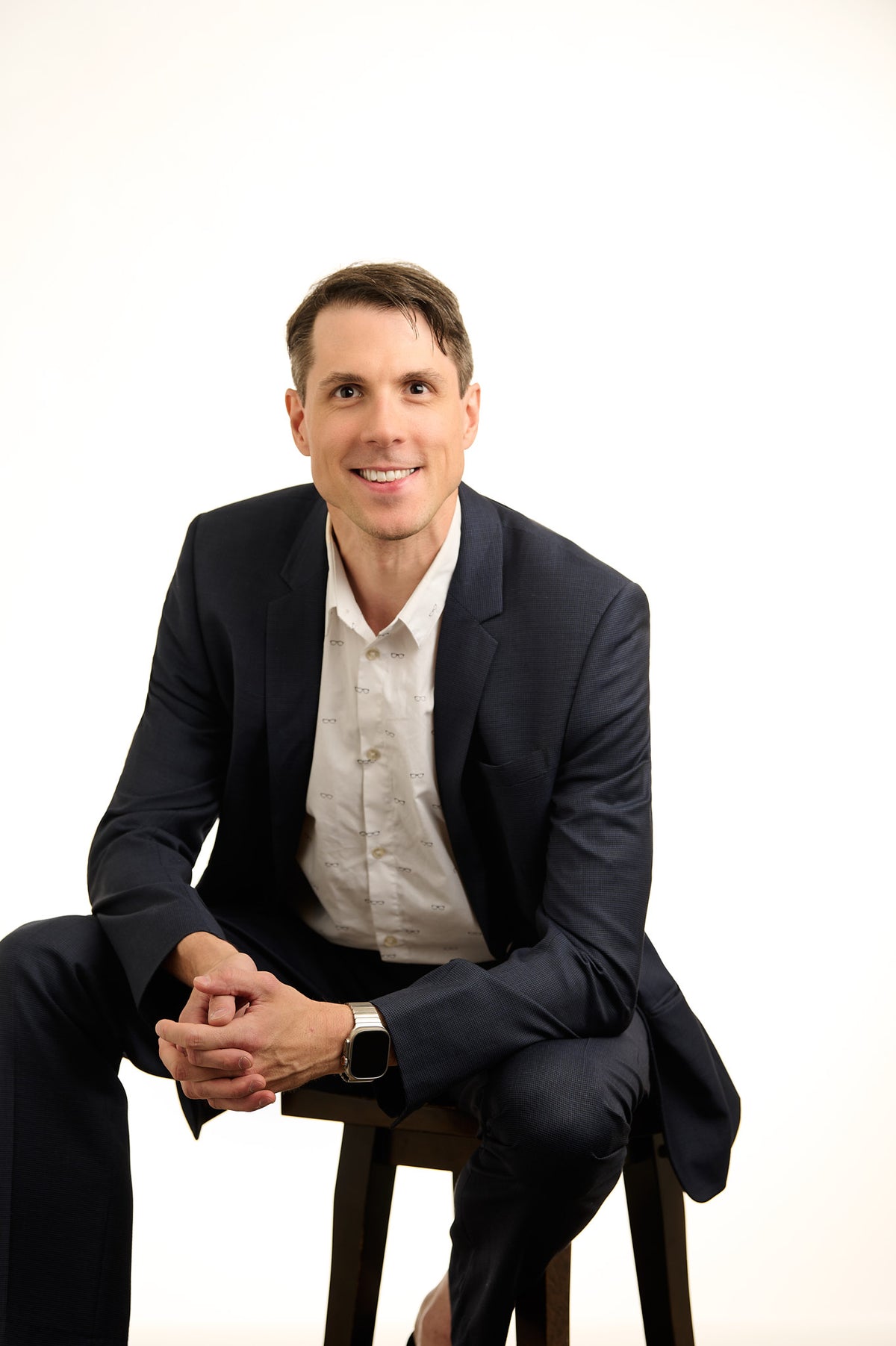 A man in a dark navy suit jacket and white button-down shirt sits on a wooden stool against a solid white studio background.