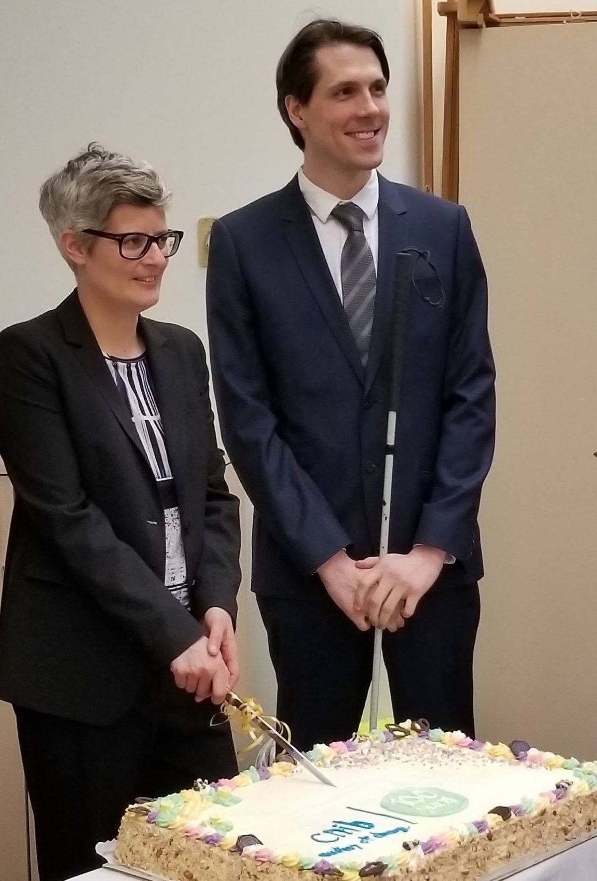 Two people standing indoors around a decorated rectangular cake on a table, with one person in a dark suit cutting the cake.