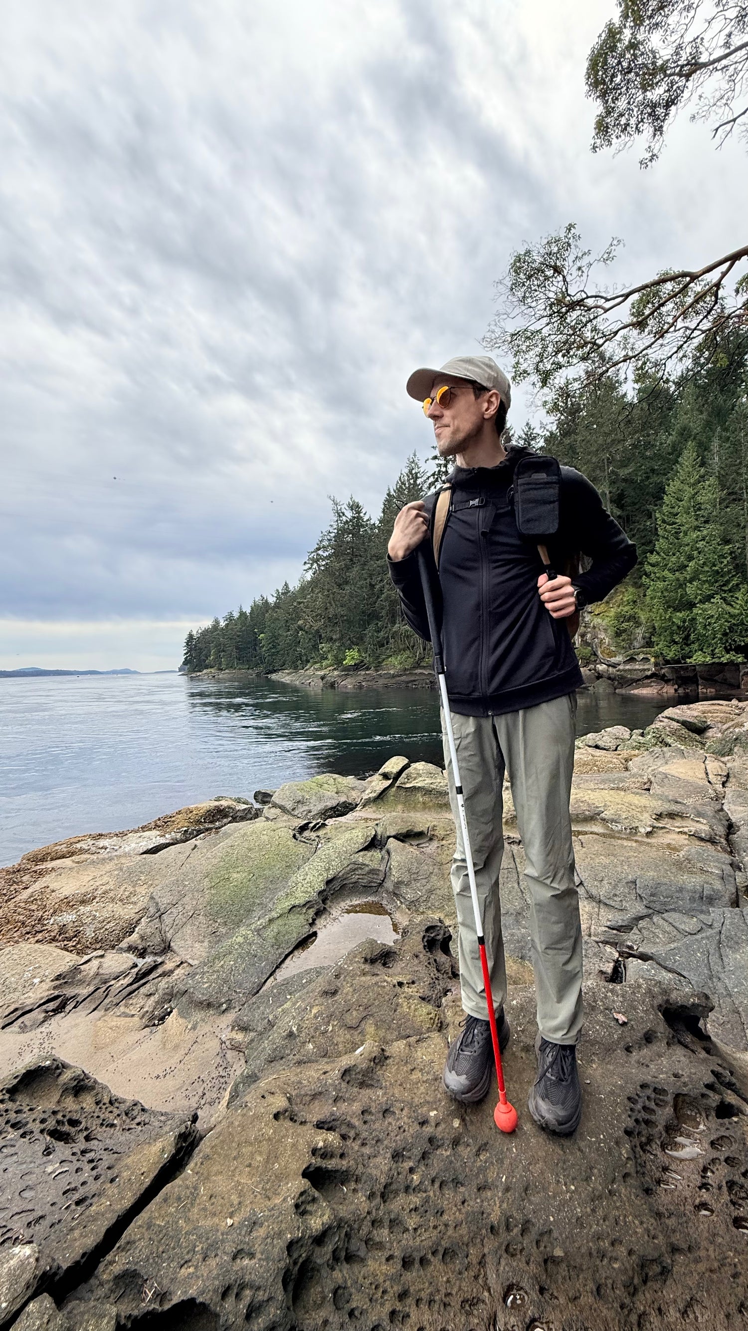 A man stands on a rocky lakeshore wearing hiking gear, including a black long-sleeve top, light olive pants, a beige cap, and sunglasses, with a backpack and trekking poles.