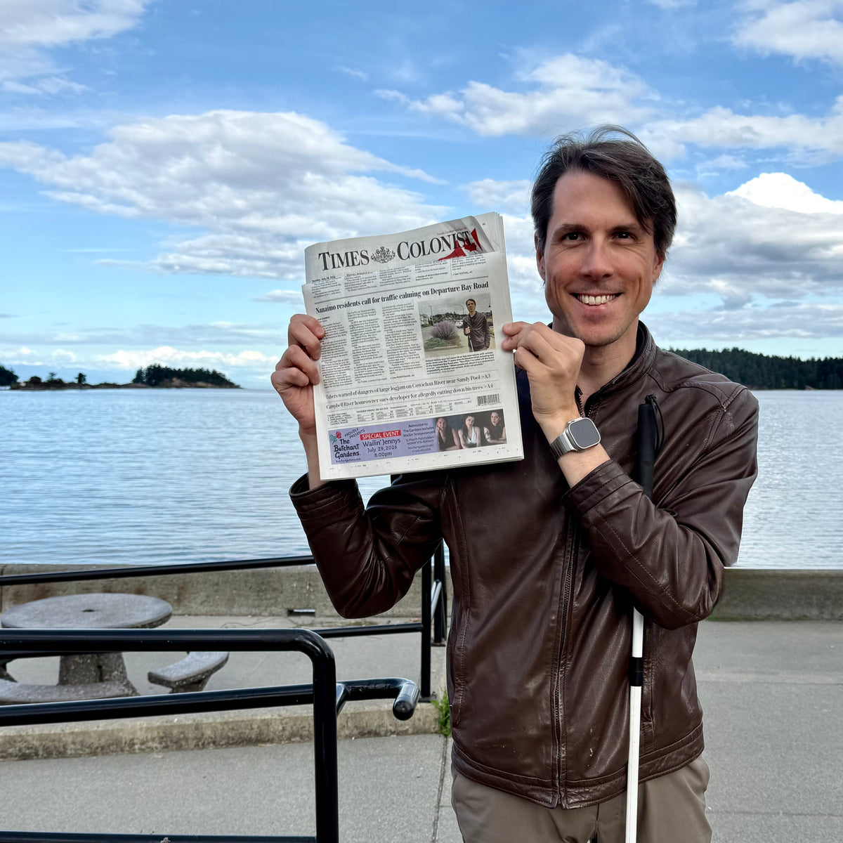 A smiling man in a brown leather jacket stands outdoors by a lake holding up a newspaper with the headline 'TIMES Colonist' visible. The background shows water, sky, distant land, and a concrete walkway.