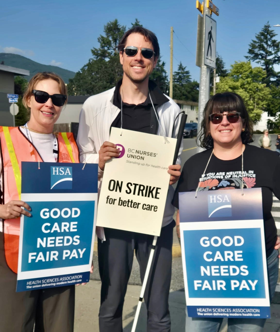 Three people standing outdoors on a street holding signs that read 'GOOD CARE NEEDS FAIR PAY' and 'ON STRIKE for better care.' Two signs also show the HSA logo.