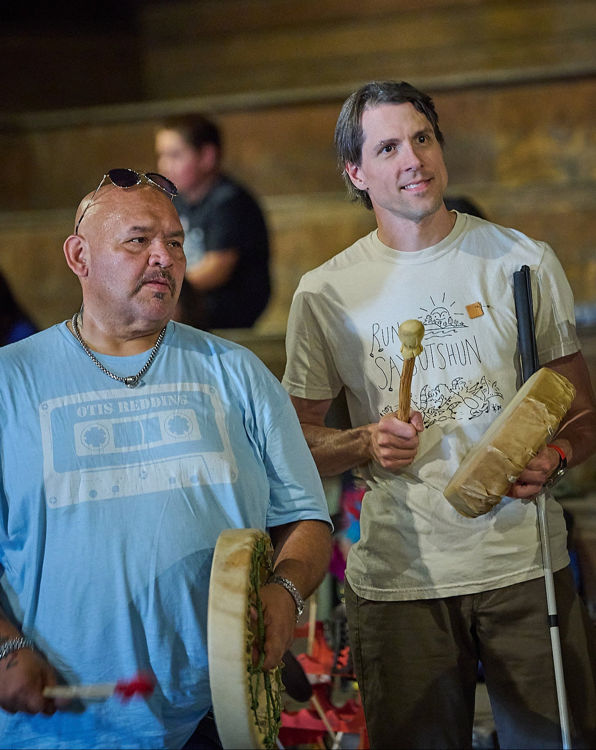 Two men standing indoors holding traditional drums and wooden mallets, wearing graphic t-shirts in a dimly lit environment.