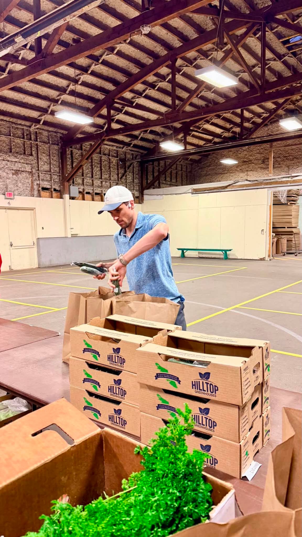 A man in a blue shirt and white cap scans a cardboard box with a handheld device inside a large warehouse or gym space. Several Hilltop branded cardboard boxes and a green leafy vegetable are visible in the foreground.