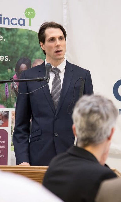 A man in a dark navy suit and tie stands at a microphone in front of a white CNIB Inca banner, speaking to an audience in an indoor event setting.