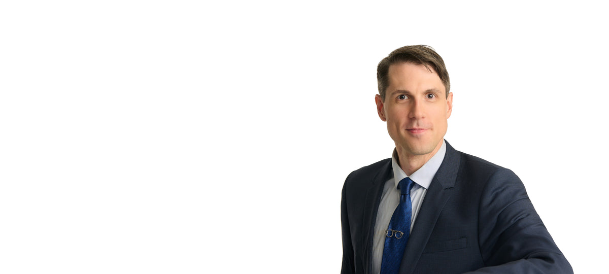A man in a dark navy suit, white shirt, and blue tie, shown from the chest up against a plain white background with his head turned to the right.