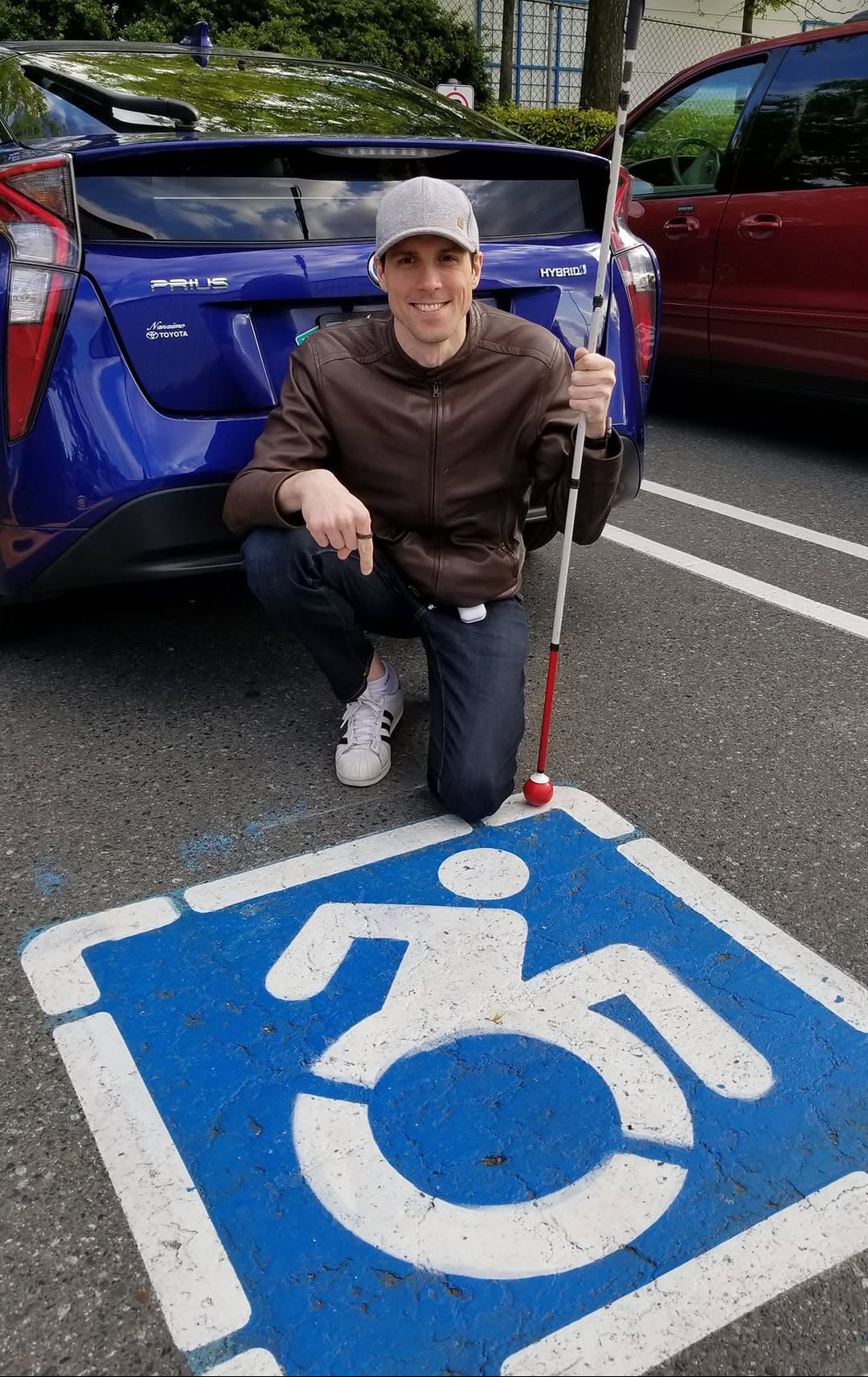A man kneels on asphalt in front of a blue car, holding a white and red mobility golf club. He is positioned over a blue and white handicap symbol painted on the ground, with other parked cars and trees in the background.