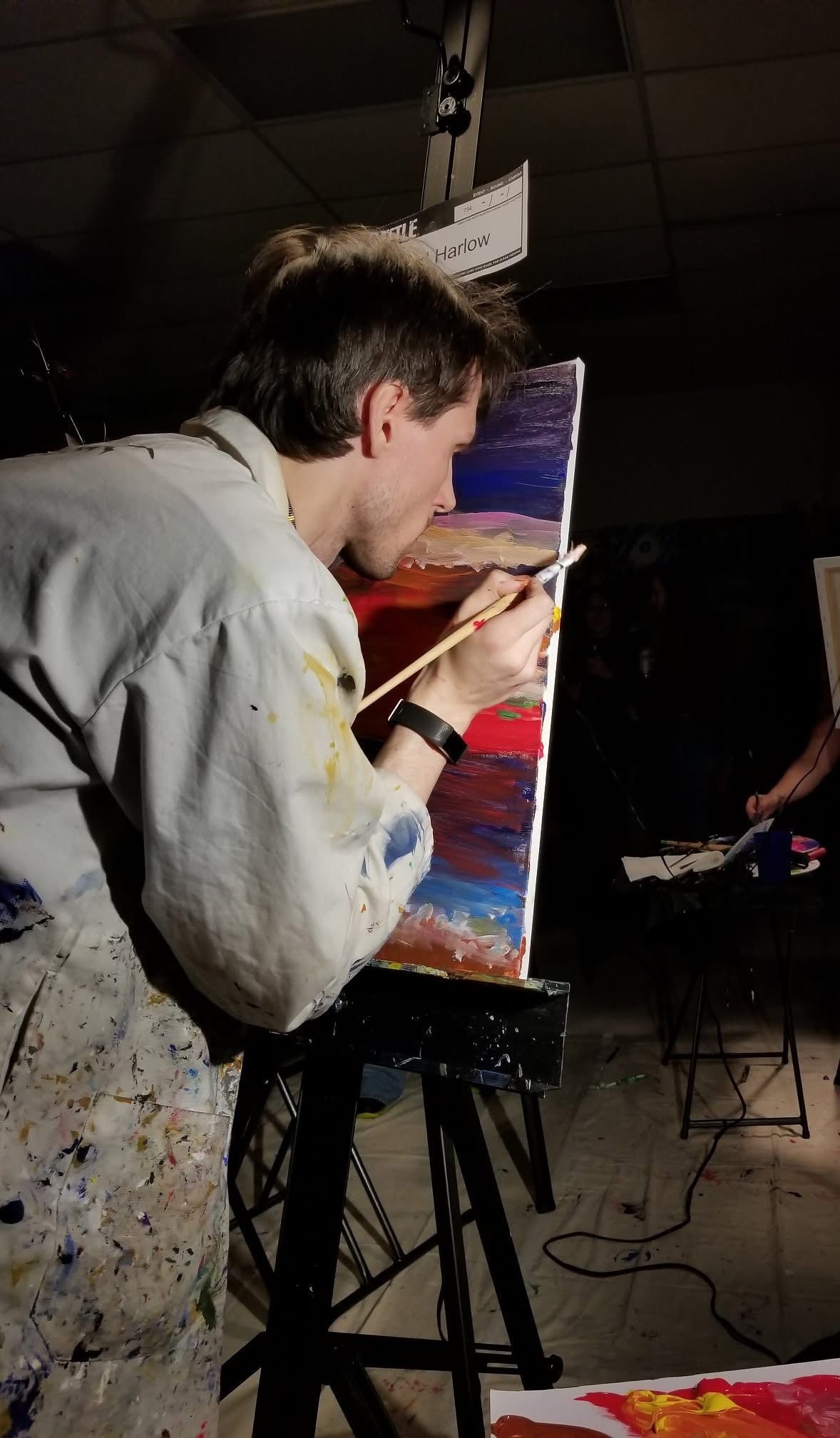 A man painting on a canvas easel in a dark studio or gallery space, wearing a light-colored painter's jacket with paint splatters on his waist. The canvas shows a colorful abstract landscape with blue, red, and yellow tones.